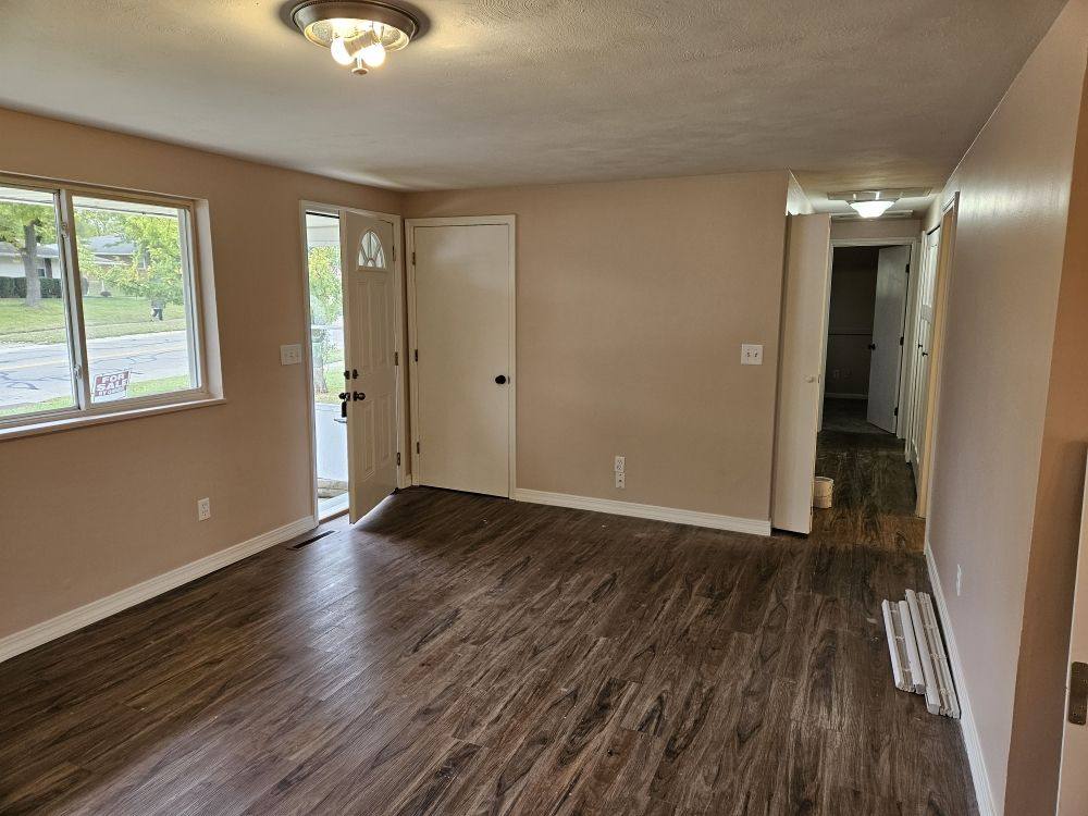 An empty freshly painted room with pale walls, white trim, a large window and new dark wood-effect flooring.