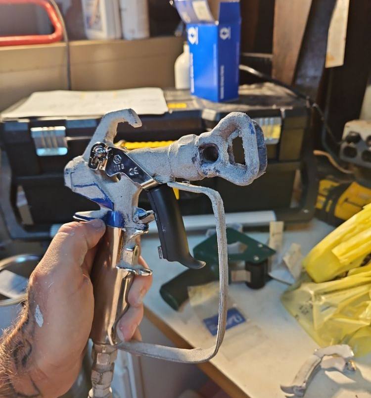 A gloved hand holding an airless paint sprayer gun over an open tool case on a job site.
